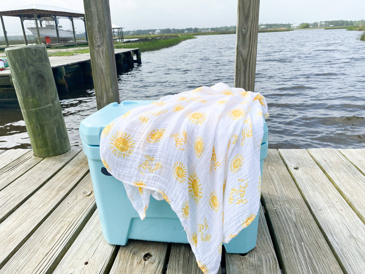 A swaddle blanket with a white base and yellow suns with Hey Y'all text all over draped over a blue cooler on a wooden dock in front of water.