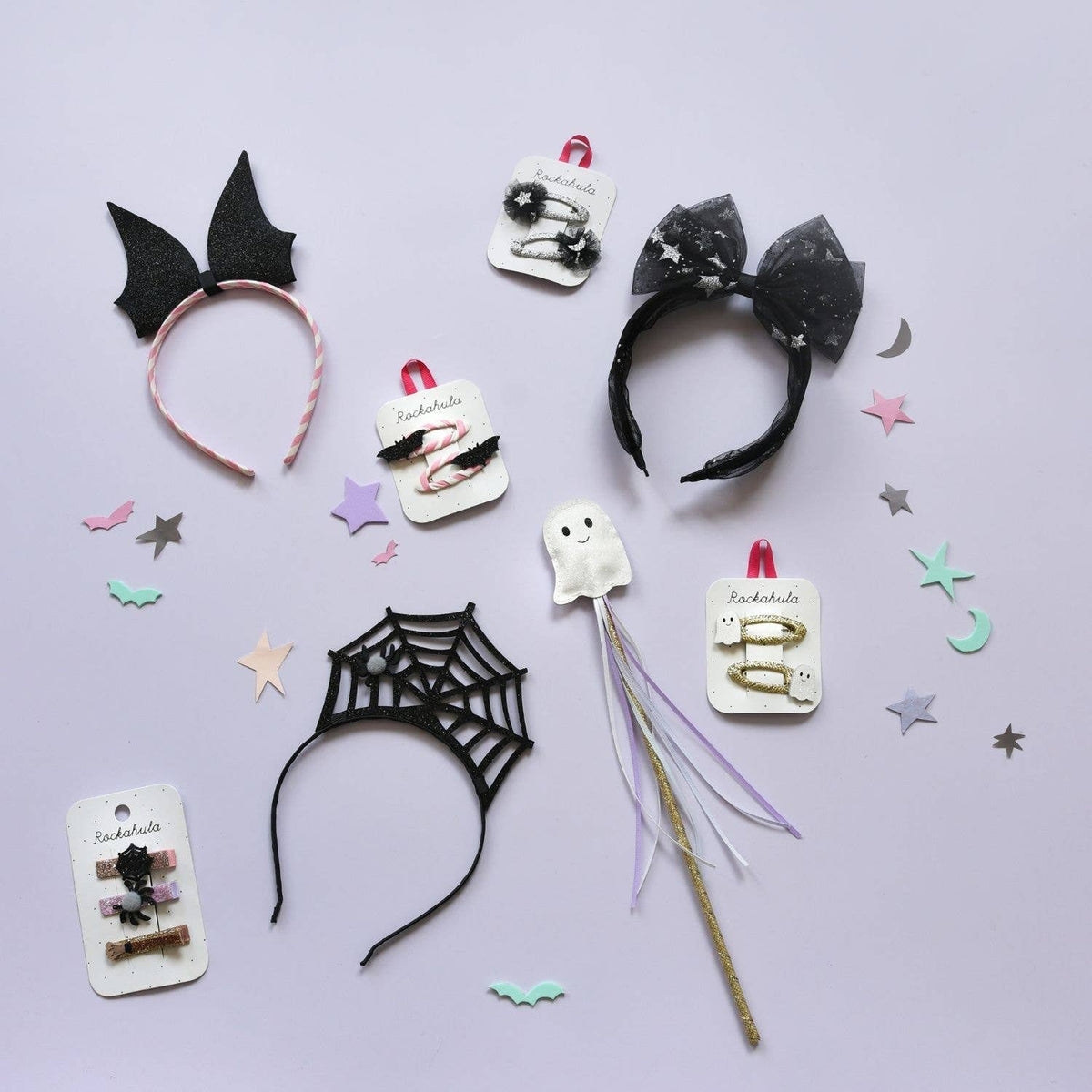 A flat-lay display of various gothic-themed hair accessories on a light lavender background, including black bat ears, a black spiderweb headband, a black bow headband with white glitter, a white ghost hair clip, and other small accessories on display cards.