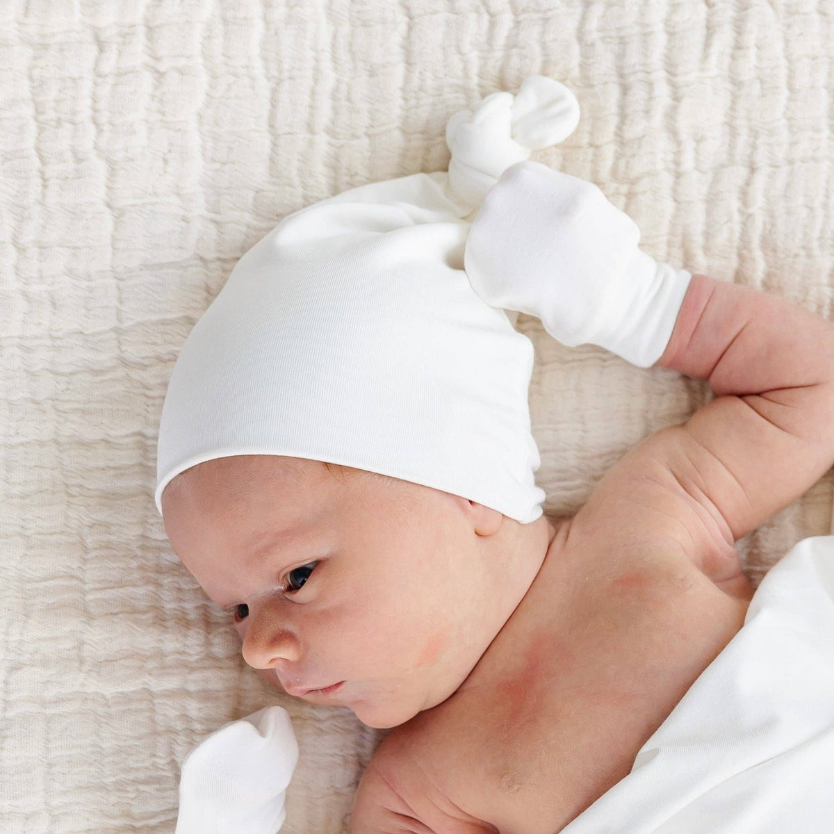 Baby is wearing a white top knot hat made of a soft fabric, designed to fit newborns to 3 months old.
