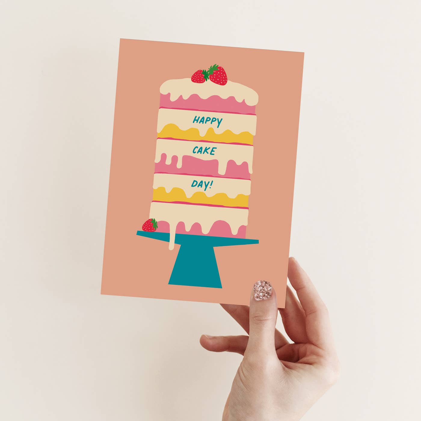 A hand with glittery nail polish holds a square greeting card featuring a colorful illustration of a three-tiered cake on a stand with the text 'HAPPY CAKE DAY!'. The card has a dusty peach background, set against a plain off-white wall.
