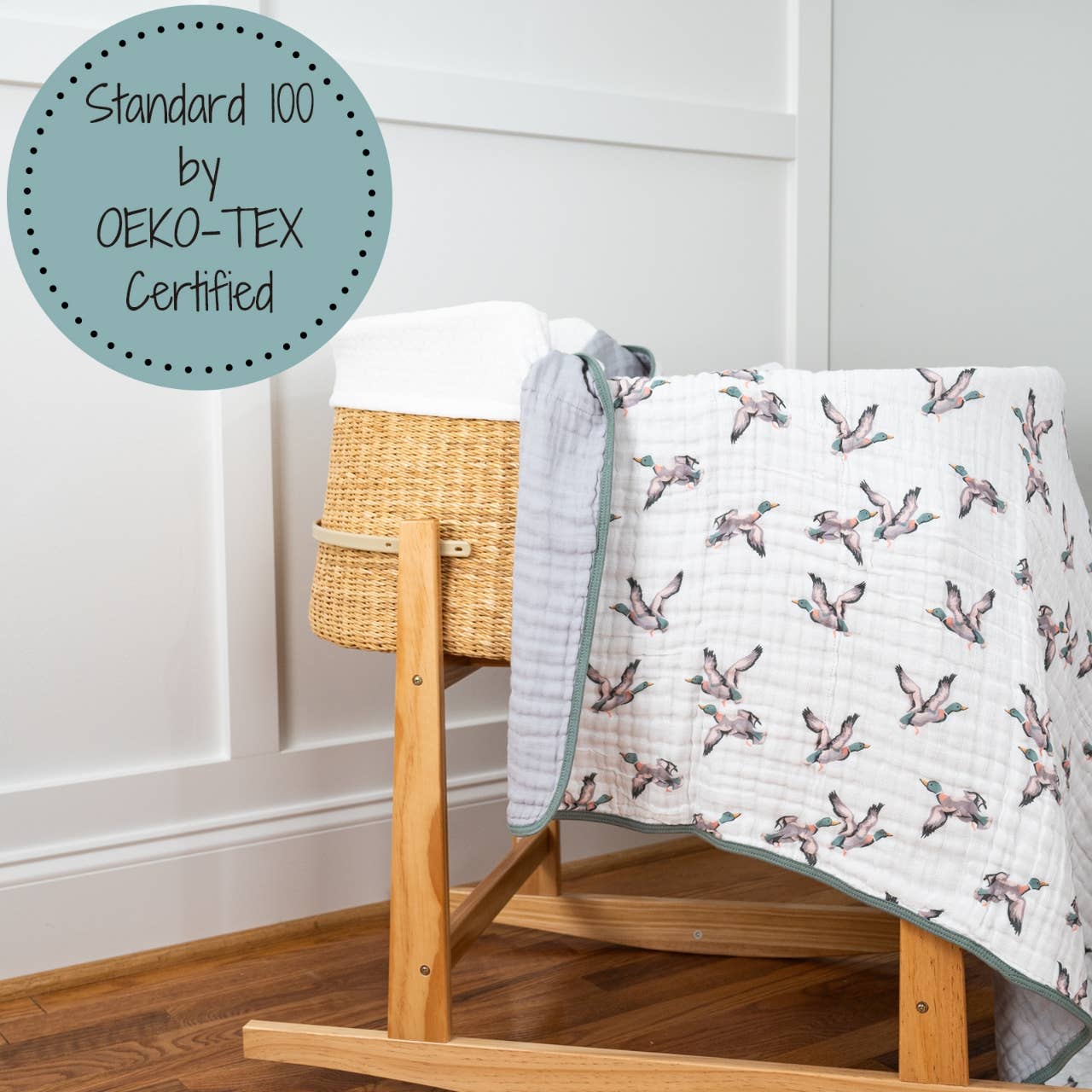A folded muslin quilt with a mallard duck print design is hanging over the side of a wooden cradle.