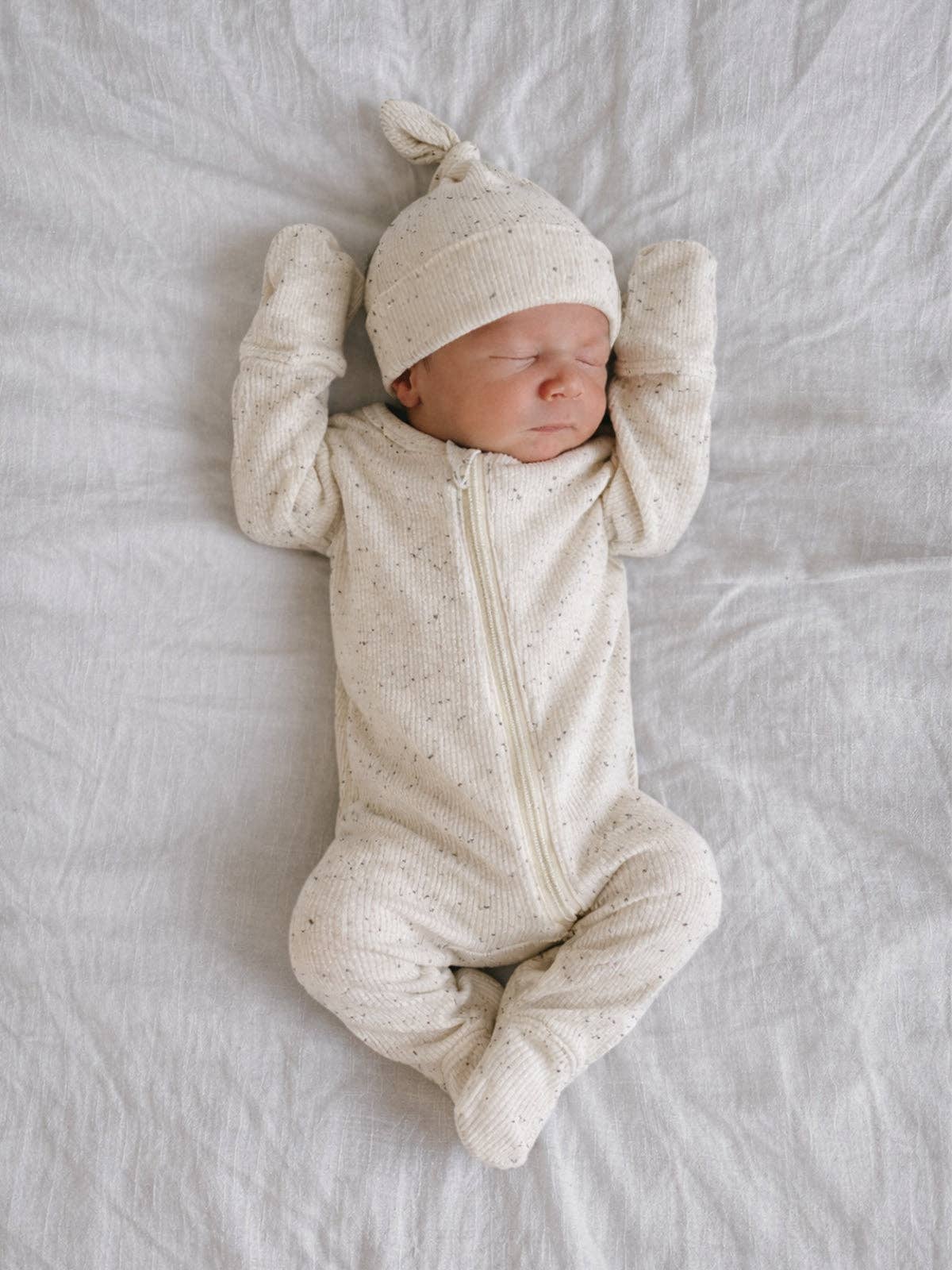 Newborn baby in a cream-colored outfit and hat lying on a soft surface