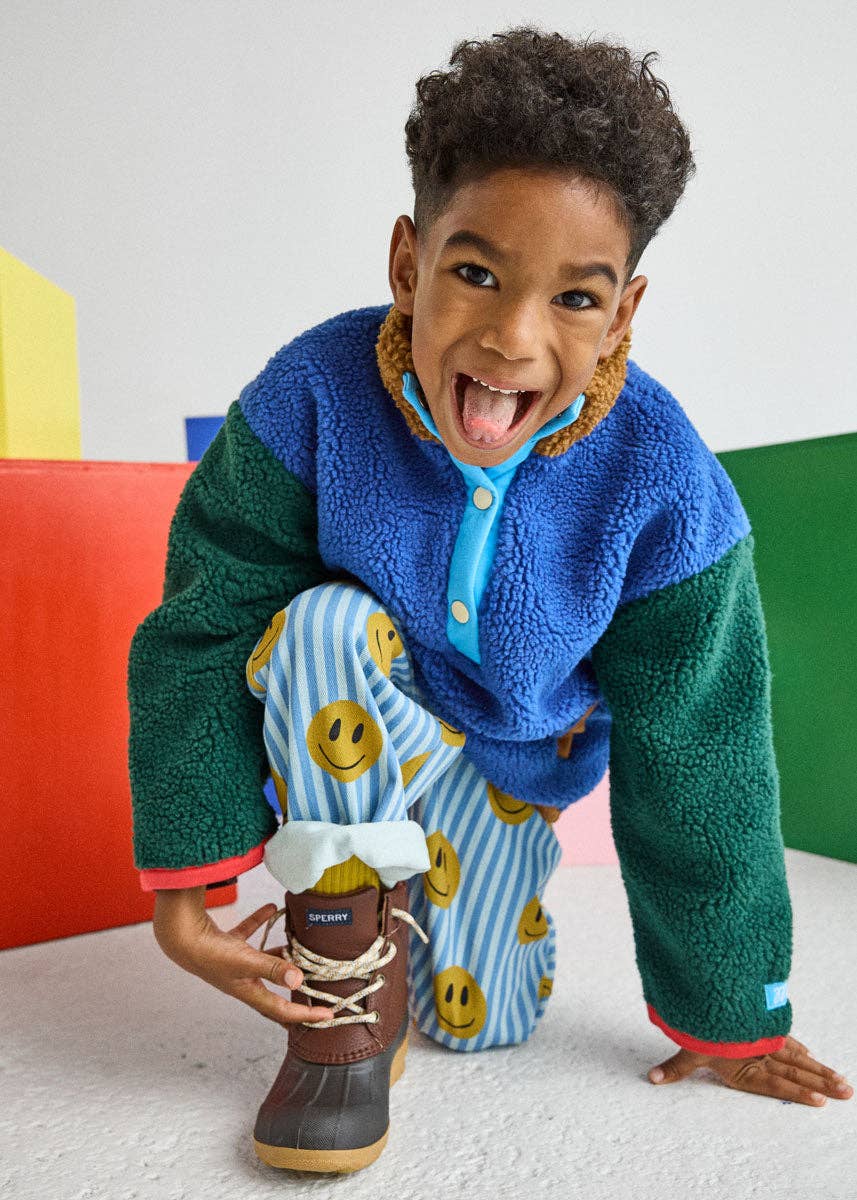A young child wearing a colorful fleece cardigan with green and blue color-blocked panels, matching striped pants with yellow smiley faces, and a brown lace-up boot, posing in a brightly lit studio with colorful geometric blocks.