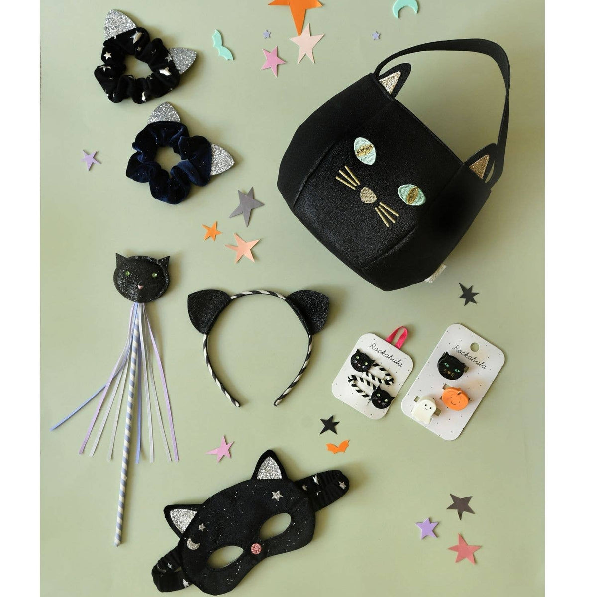Black cat-themed accessories including a bag, mask, headband, and hair ties on a green background with star decorations.