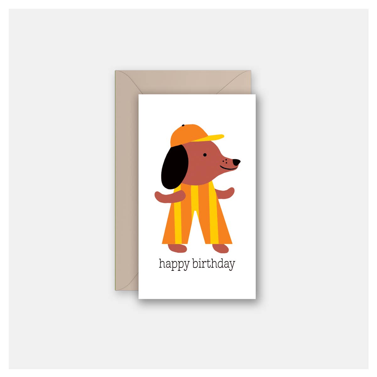 A birthday greeting card with a white front featuring a cartoon dog in an orange and yellow striped outfit and cap, with the text 'happy birthday' below. The card is shown with a plain brown envelope behind it on a light gray background.
