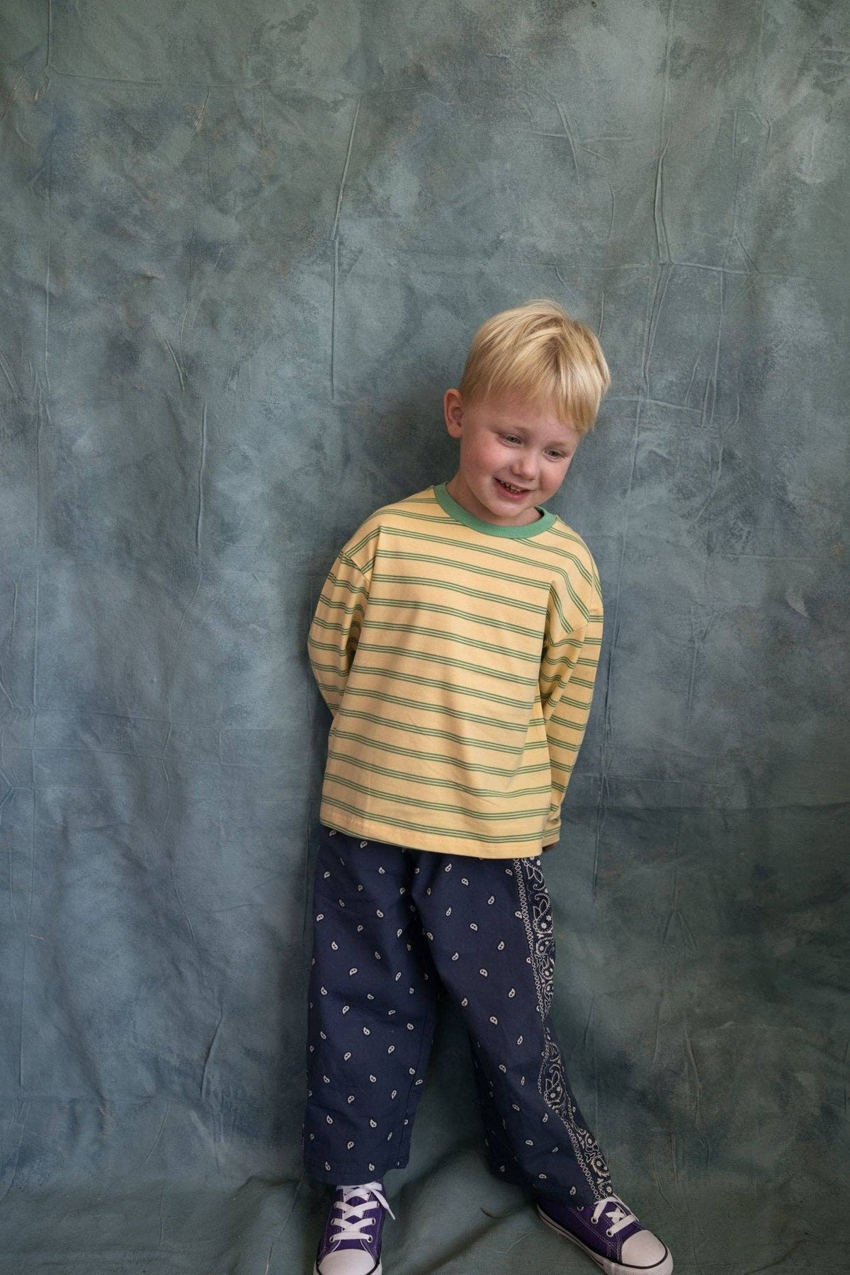 A young blonde boy wearing a yellow and green striped long-sleeve shirt, navy pants with white star patterns, and purple sneakers, standing against a mottled blue-gray studio backdrop.