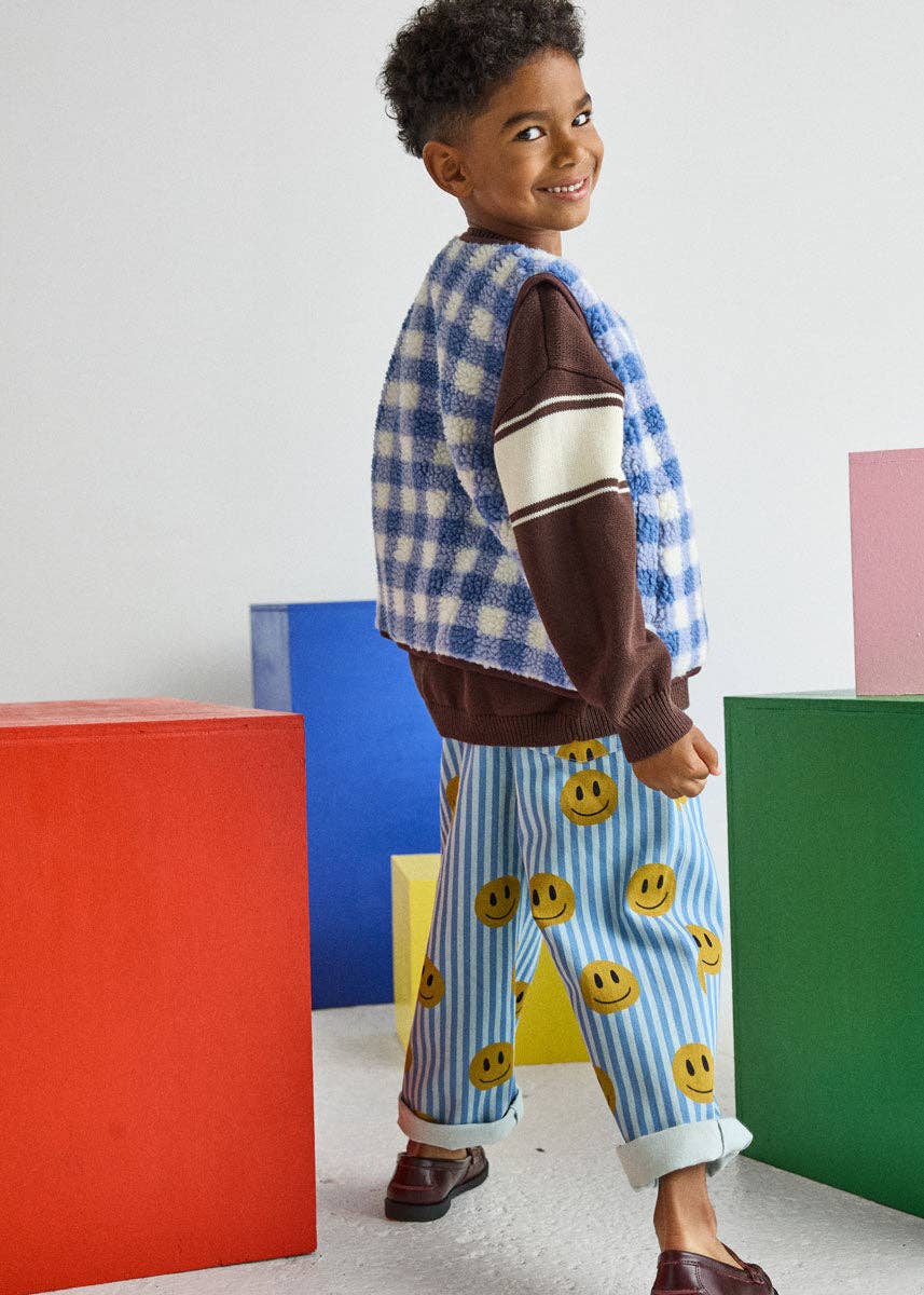 A young boy wearing a blue and white gingham vest over a brown long-sleeve shirt with white stripes, paired with blue striped pants featuring yellow smiley faces. He is standing in a studio setting with colorful geometric blocks in the background.