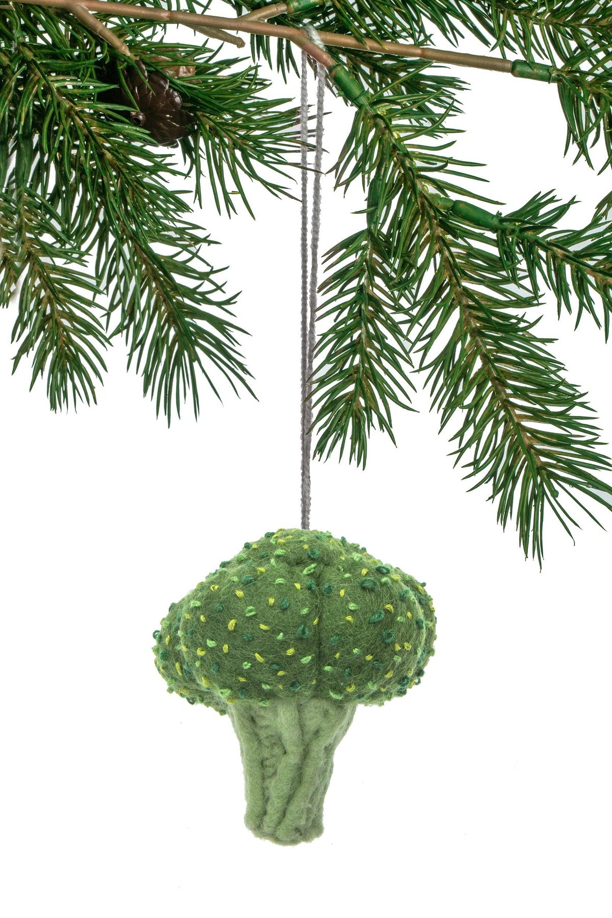 A green felt broccoli Christmas ornament hanging from a silver string beneath pine tree branches against a white background.