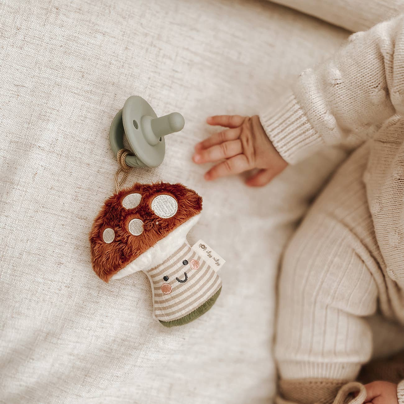 A plush mushroom lovey with a pacifier attached to it, designed for babies on a tan surface next to a baby.