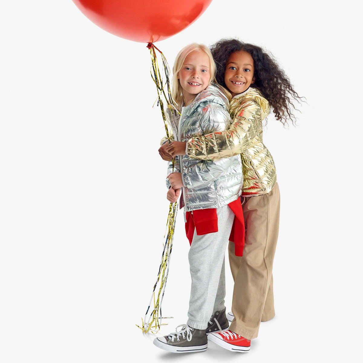 Two children hugging while wearing shiny metallic costumes, one in a silver jacket and the other in a gold jacket, holding a large red balloon with tinsel against a light gray background.