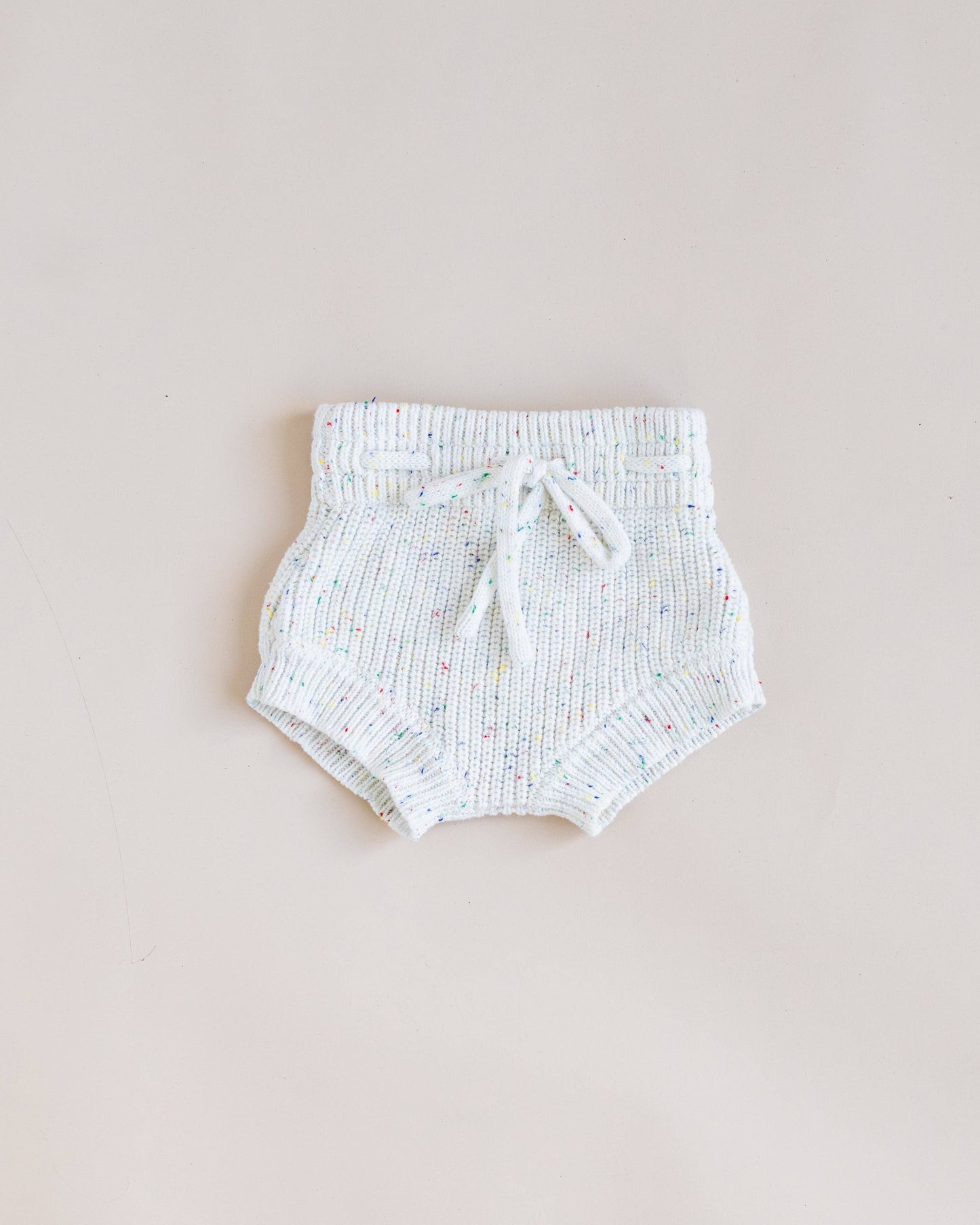 A pair of white knit baby bloomers with a textured ribbed pattern and a matching drawstring tie, laid flat on a light beige background.
