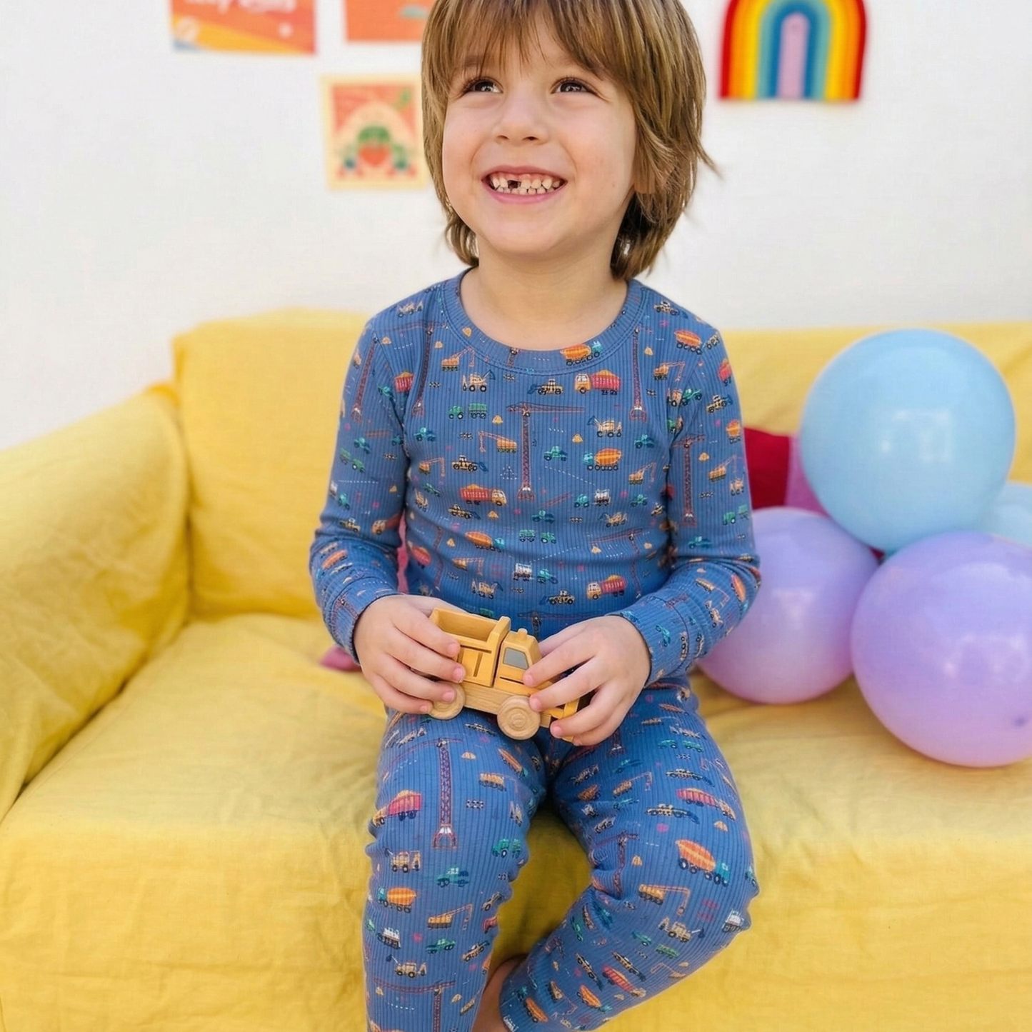 Child in blue pajamas with car pattern sitting on a yellow couch with balloons.