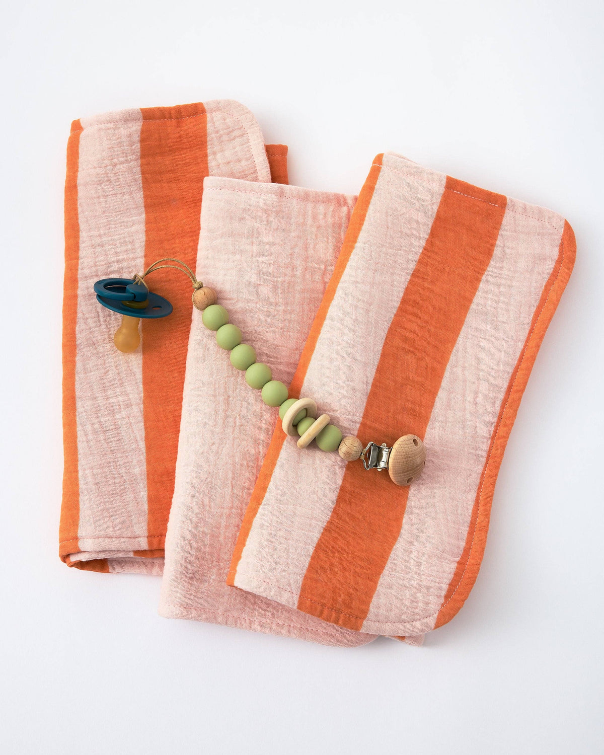 Three folded striped muslin burp cloth in orange and pale pink, styled with a wooden bead pacifier clip on a white background.