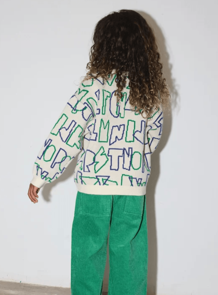 A young child with curly dark hair stands indoors. The child is wearing a white long-sleeve sweater covered in green and blue graffiti-style lettering and a bright green corduroy pants.