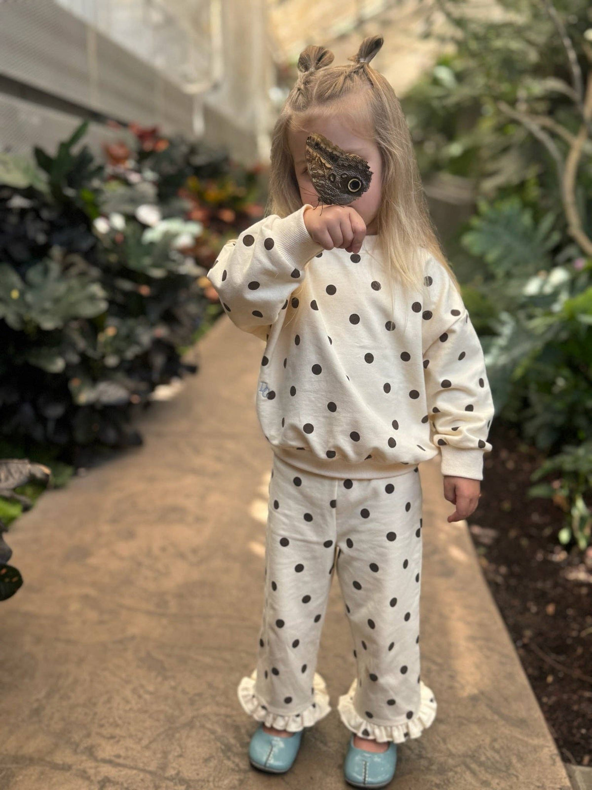 A young child wearing a matching cream-colored pajama set with black polka dots and ruffled hems, standing on a stone path in a garden.