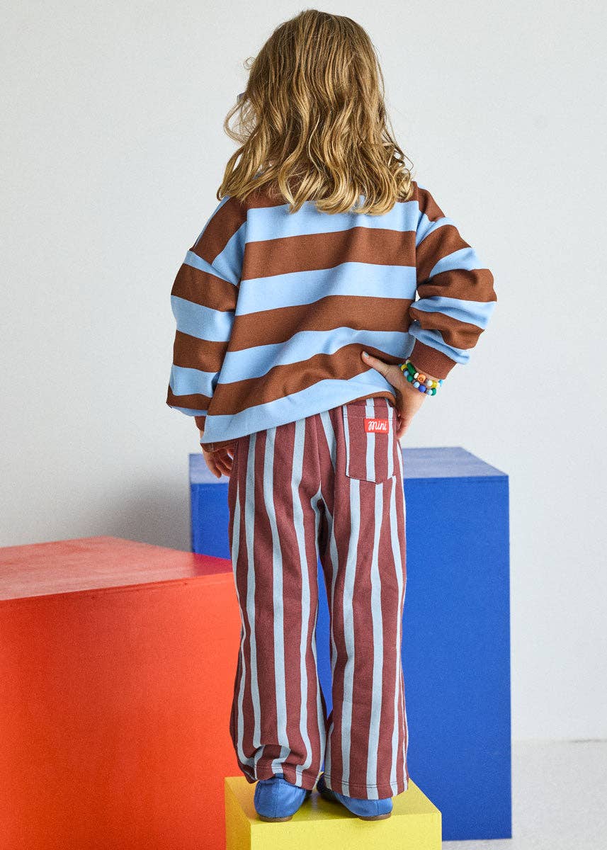 A child standing with their back to the camera, wearing a brown and light blue horizontally striped long-sleeve top and matching brown and white vertically striped pants, posed on colorful geometric blocks against a white wall.