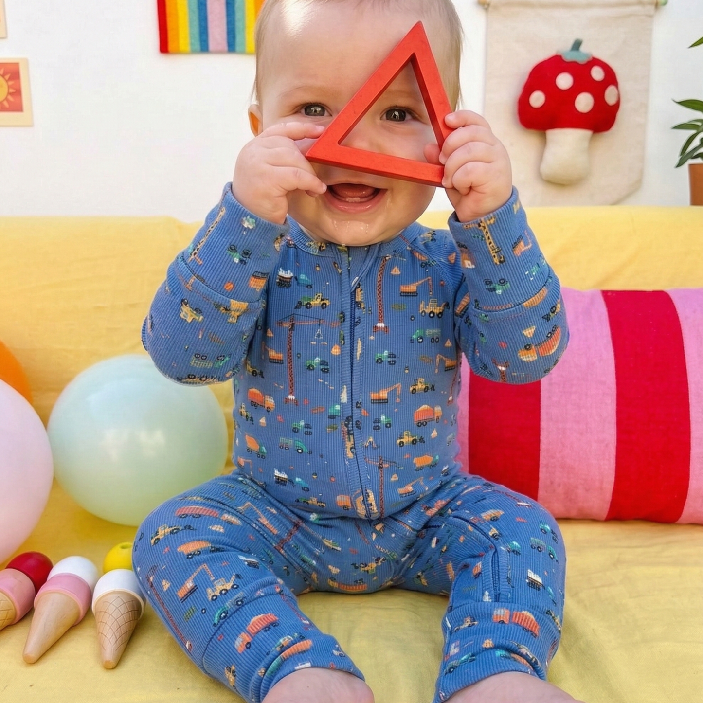 Baby wearing a blue onesie with vehicle patterns, holding a red triangle to their face, sitting on a colorful couch.