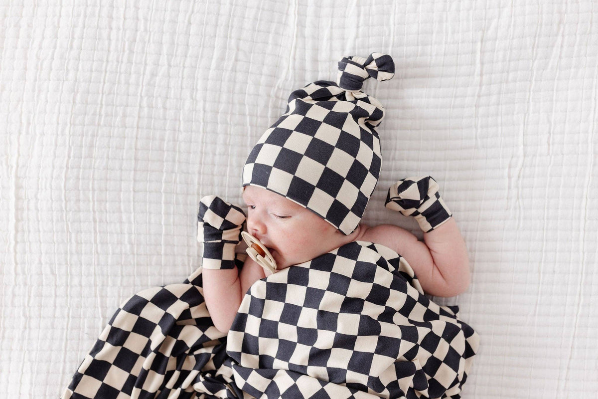 Baby is lying on a textured white background and wearing being and black checkerboard top knot hat, no scratch mittens and wrapped in a checkerboard patterned blanket.