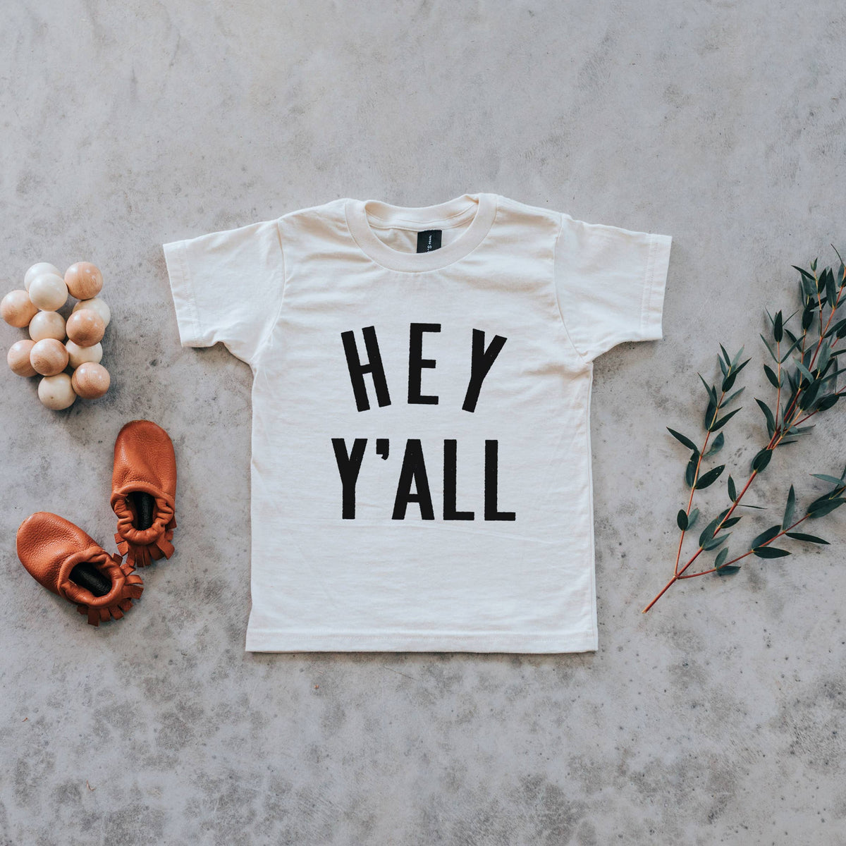 A white crew neck t-shirt with the text 'HEY Y'ALL' printed in black, displayed on a concrete background accompanied by wooden eggs and a small branch.