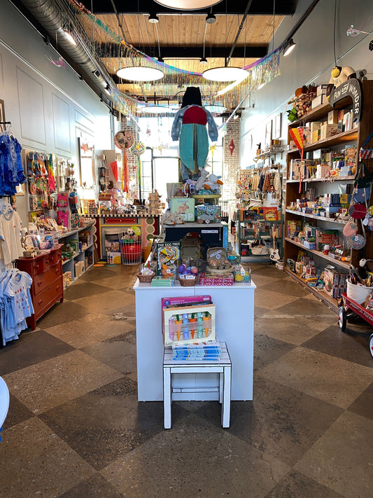 Photo of a retail store showing a stool with books, clothing hangong on pegs, stacks of books and bookshelves with toys and games.