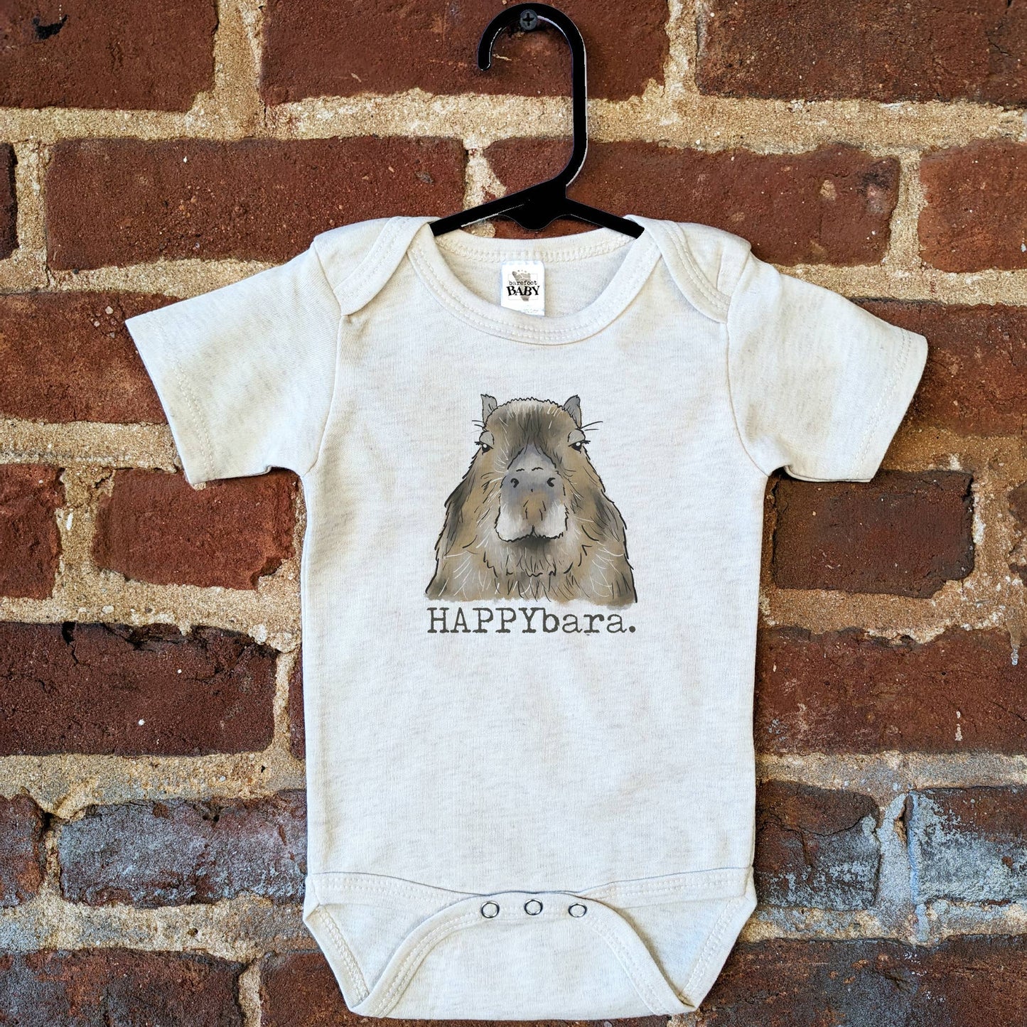 Baby onesie with capybara design and 'HAPPYbara' text on a brick wall background