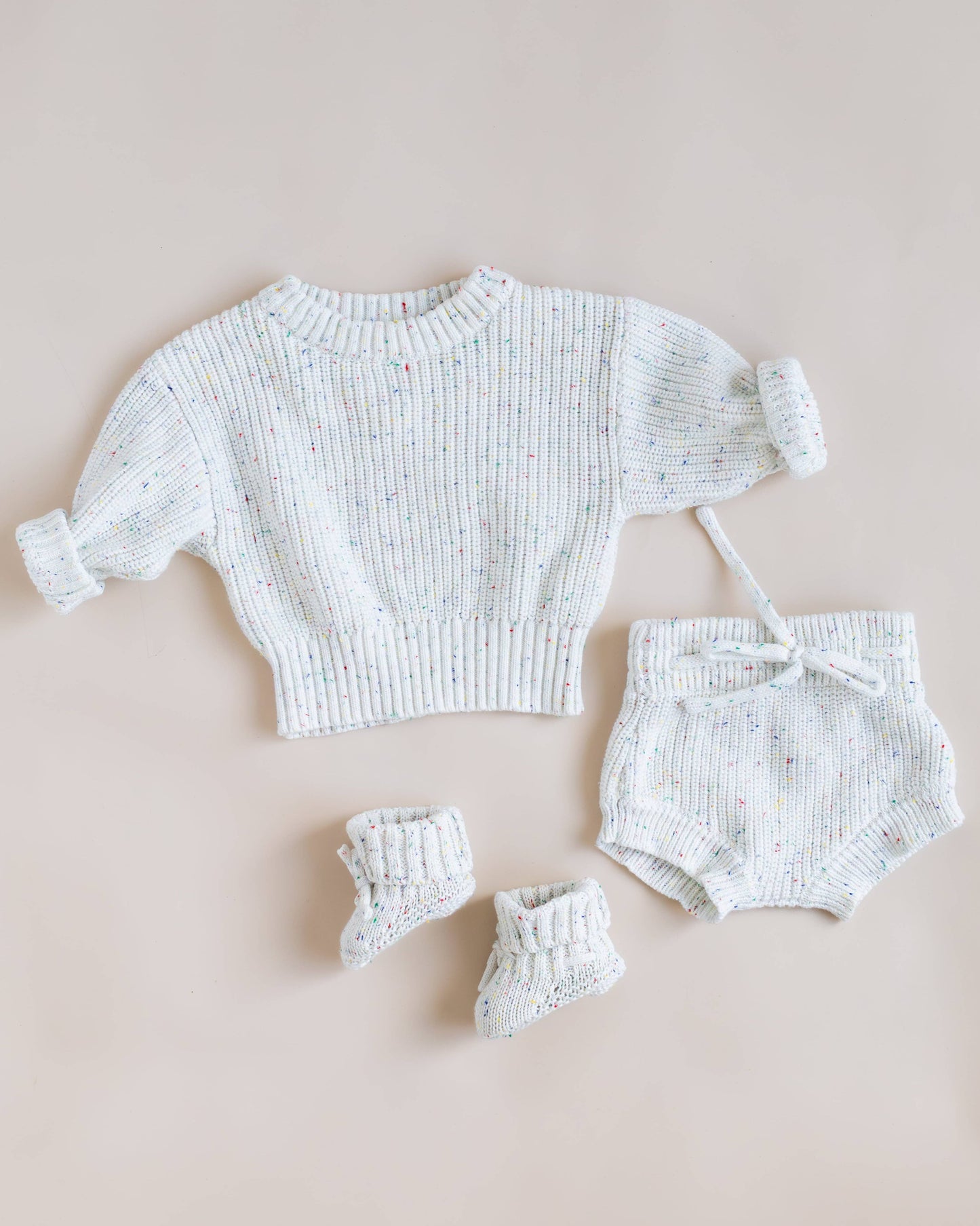 A baby knit clothing set laid flat on a soft beige background, including a short-sleeve ribbed knit top, matching bloomers, and booties in a very light off-white or pale blue-gray yarn with tiny multicolor flecks.