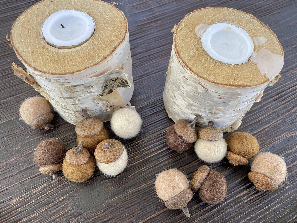 Two natural birch log candle holders with white wax inserts, surrounded by small acorns and woolen decorative accents on a dark wood surface.