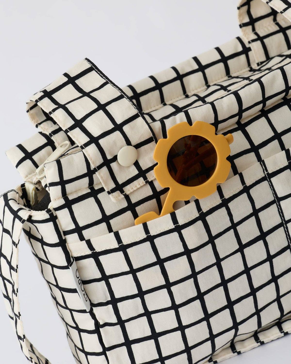 A close-up of a cream canvas shoulder bag with a black grid pattern, featuring a side pocket with a yellow sun-shaped sunglasses holder and a pair of yellow sunglasses in it.