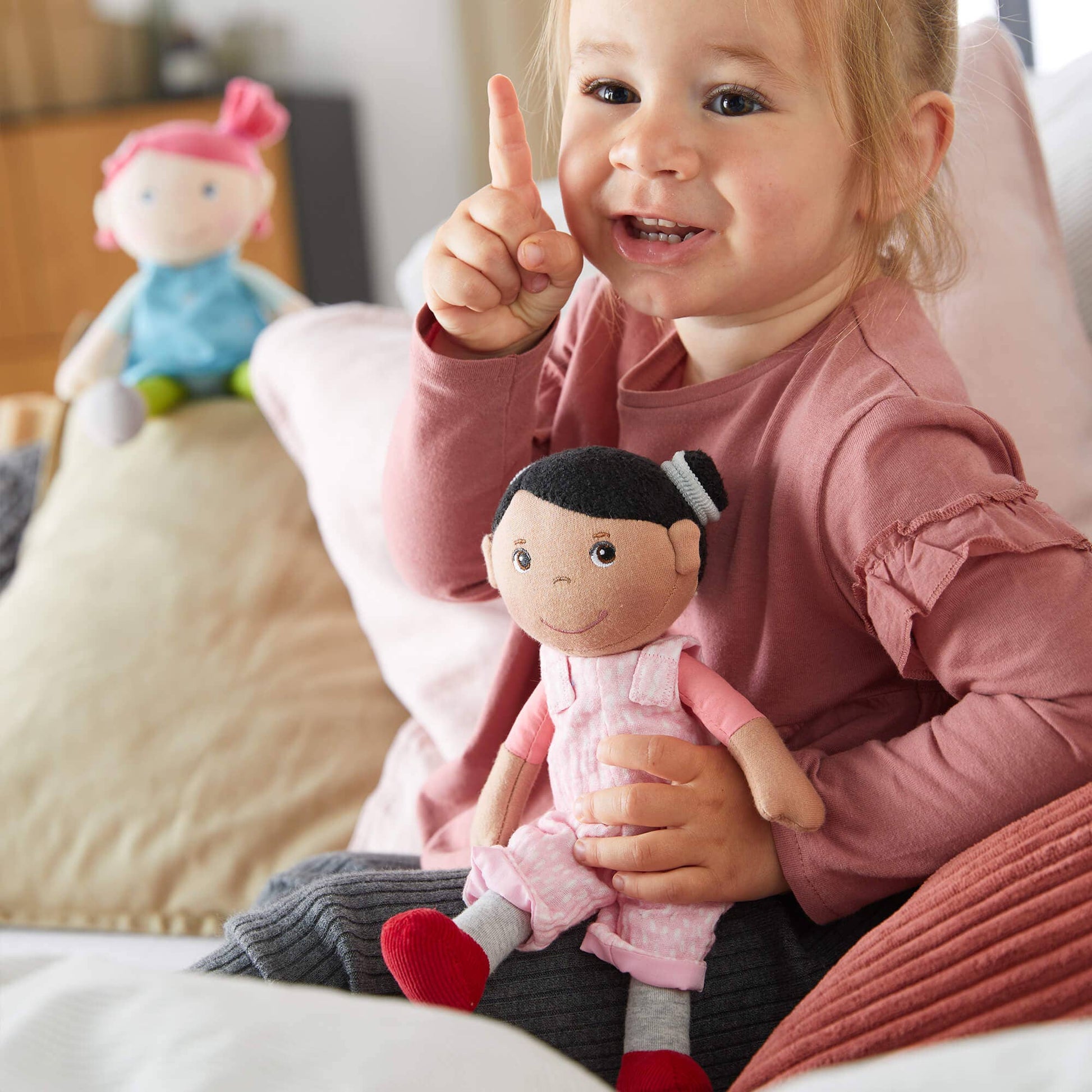 Child holding a doll and pointing upwards with a smile, with another doll in the background.
