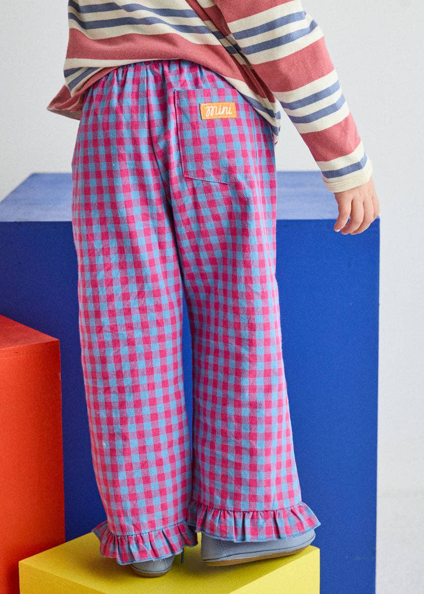 A child wearing pink and blue gingham wide-leg pants with ruffled hems, paired with a striped long-sleeve top and gray slip-on shoes, standing on colorful geometric blocks.