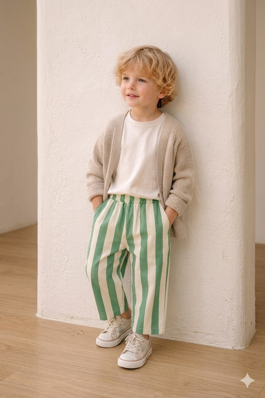 Child wearing a beige sweater, white shirt, and green striped pants standing against a white wall.