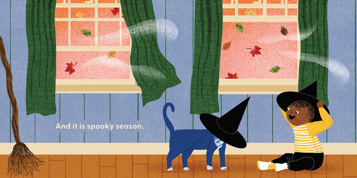 Illustrated children's scene showing a child in a yellow striped shirt and black witch hat sitting on the floor, looking at a blue cat wearing a black witch hat. Two windows with green curtains and falling leaves are visible in the background, with the text 'And it is spooky season.