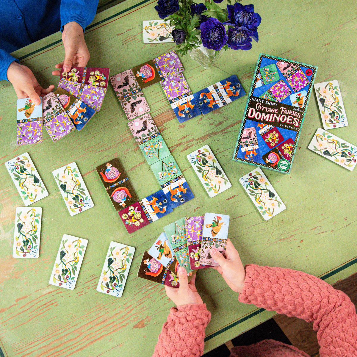 Colorful dominoes game being played on a green table with hands interacting with the tiles.