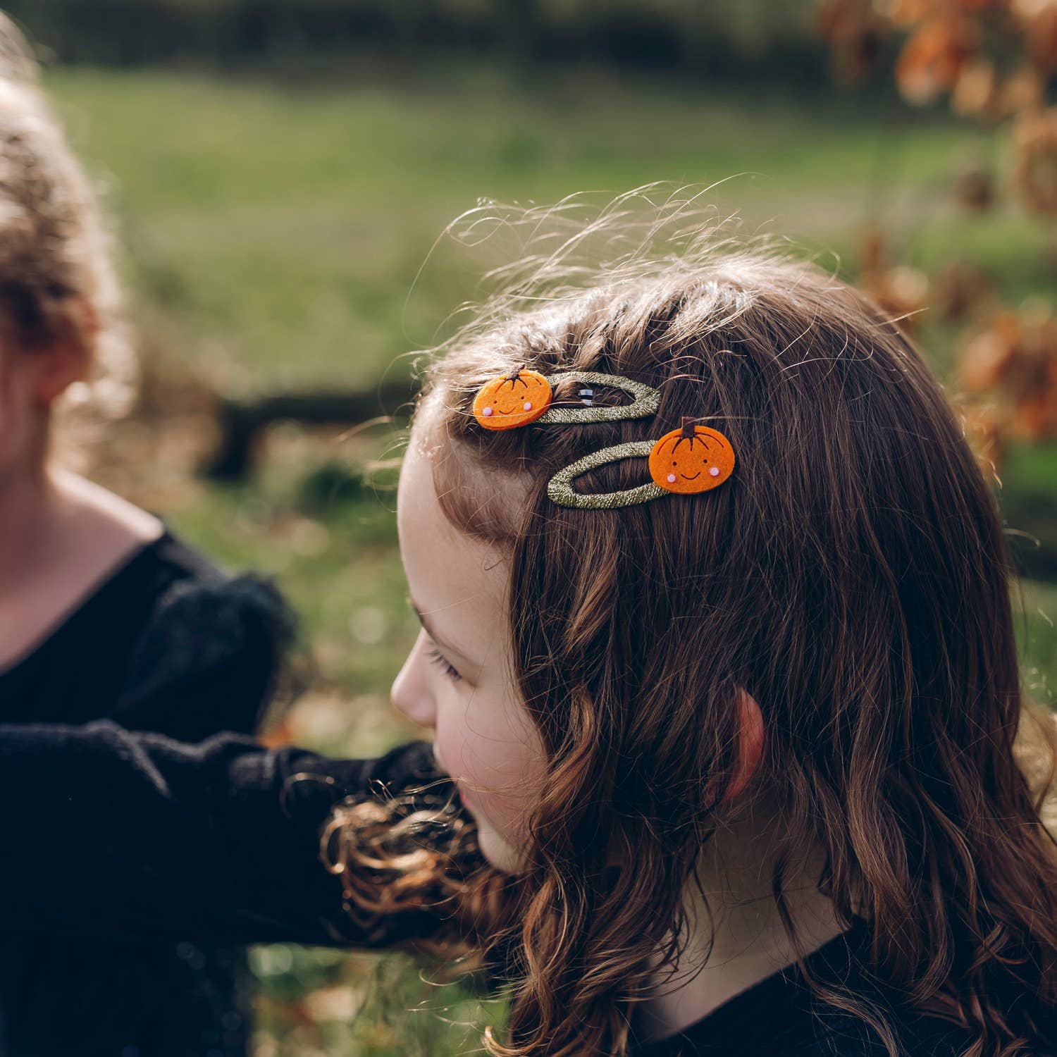 A close-up lifestyle shot of a young girl with brown hair wearing three pumpkin-shaped hair clips with silver glittery bands, set against a blurred outdoor background.