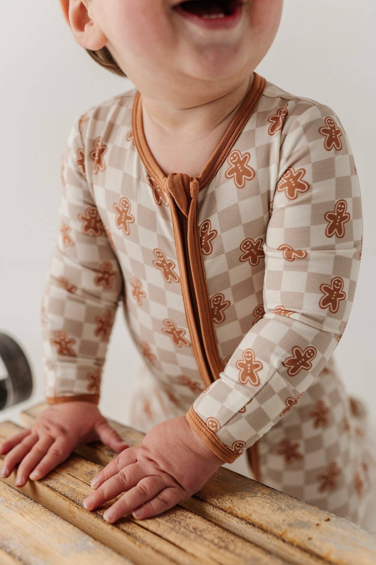 A close-up of a baby wearing a long-sleeve one-piece romper with a beige and white checkerboard pattern and brown gingerbread cookie motifs. The romper features a brown zipper and trim, and the baby is leaning on a rustic wooden surface.