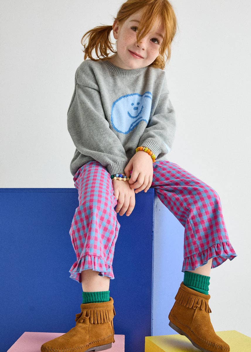 A young girl with red hair sits on a colorful blue and yellow block, wearing a gray sweater with a blue smiley face, pink and purple gingham pants, green socks, and brown suede boots.