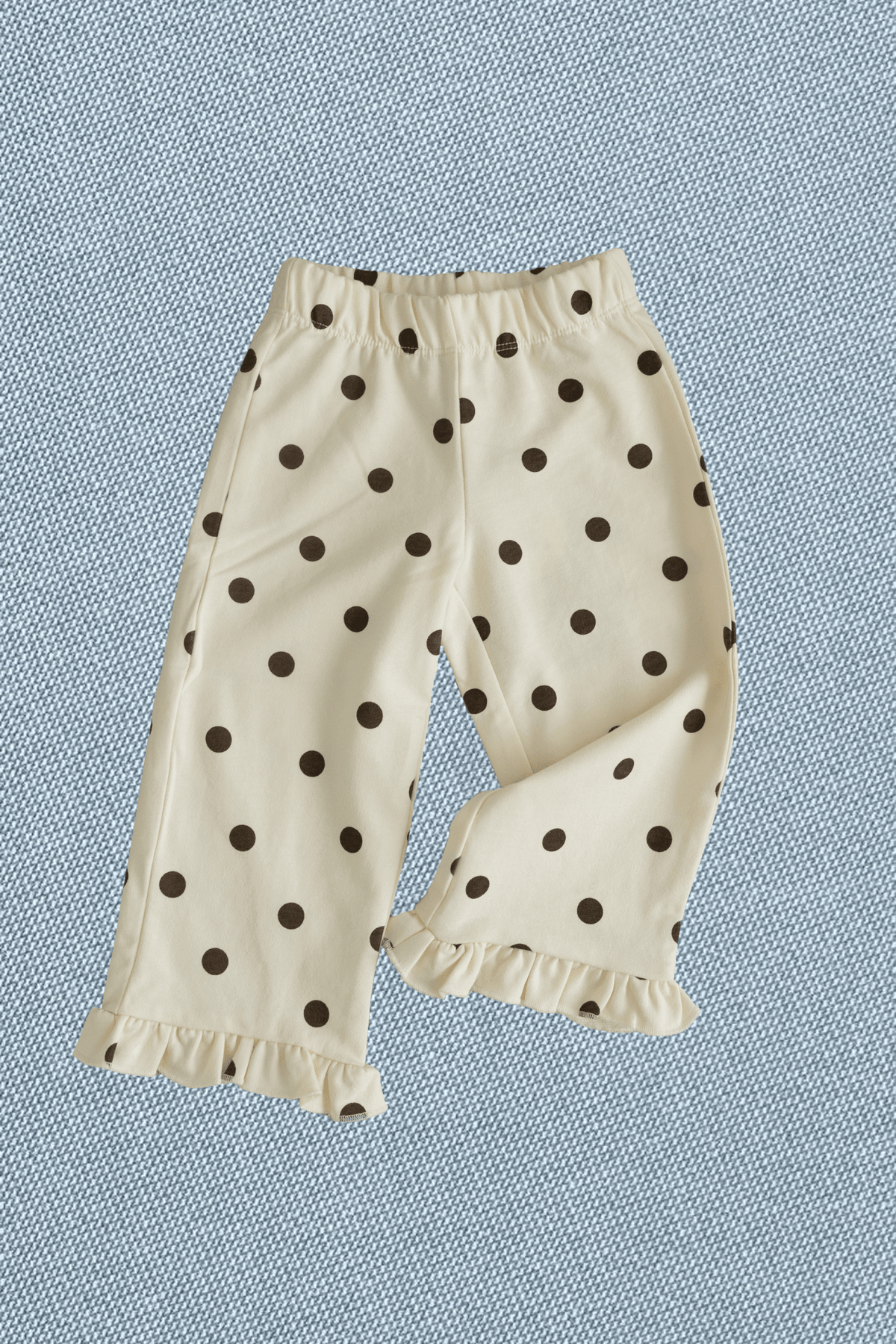 Cream-colored toddler pants with a dark brown polka dot pattern and ruffled hems, laid flat on a textured blue background.