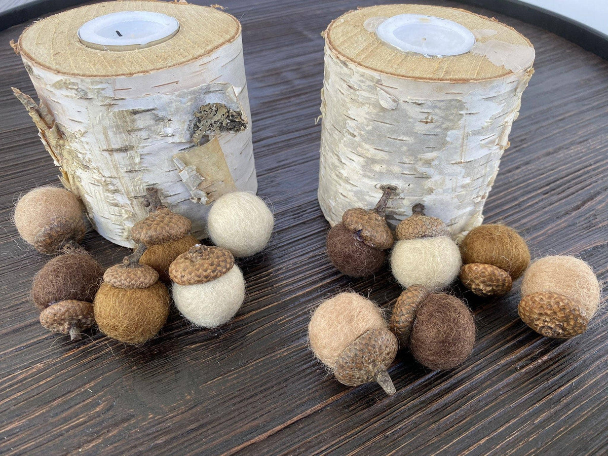 Two birch log candle holders with white tea light inserts and natural wood tops, arranged on a dark wooden surface with felt acorns in brown, tan, and cream tones.