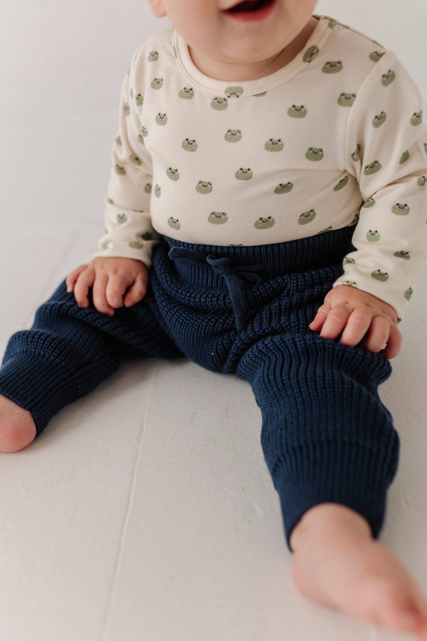 Baby wearing a white long-sleeve shirt with green patterns and dark blue knitted pants on a light background