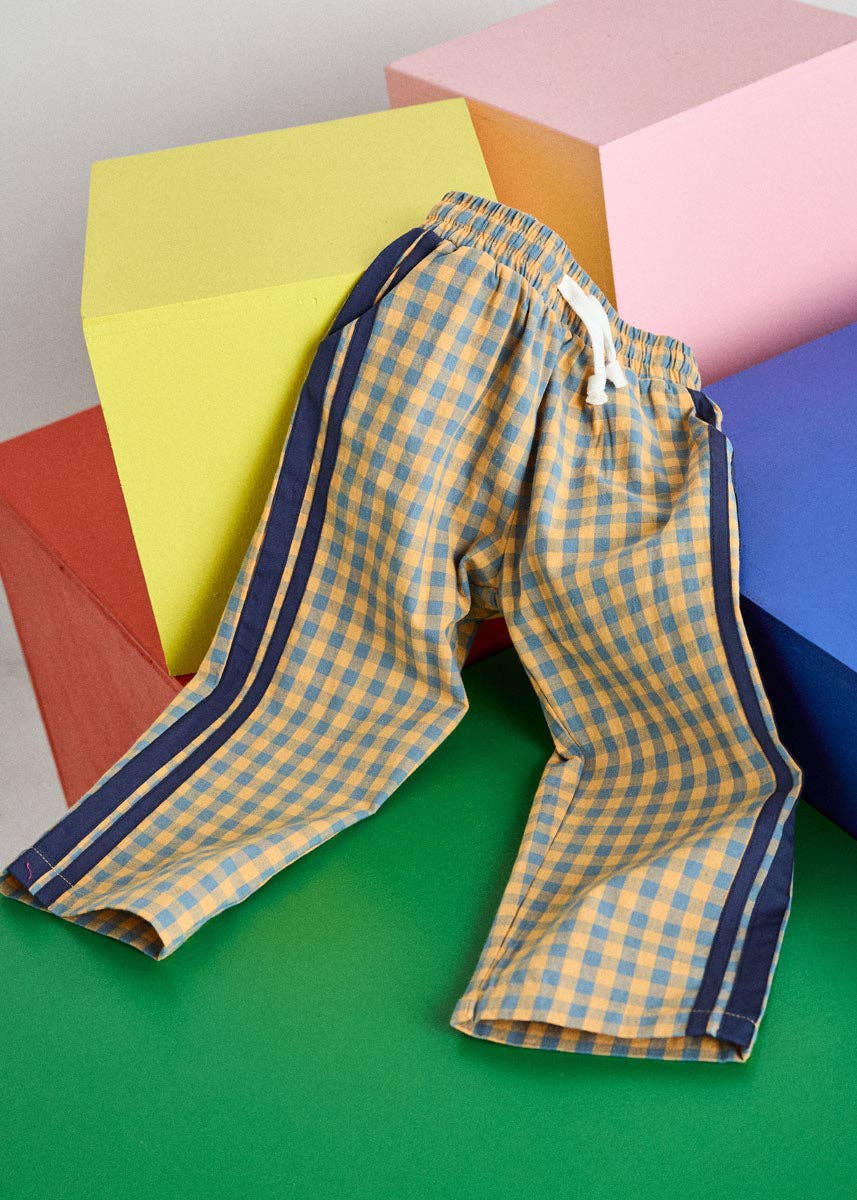 A pair of yellow and blue gingham check pajama pants with navy blue side stripes, styled on a green surface with colorful geometric blocks in the background.