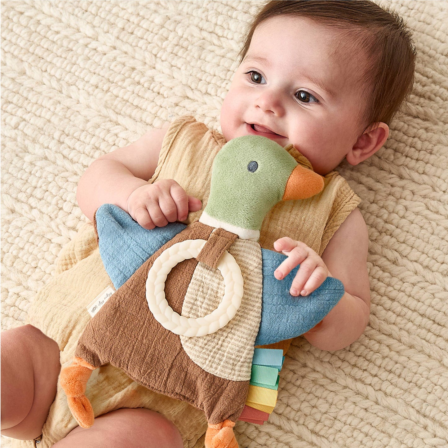 A baby lying on a textured cream blanket, smiling while holding a plush duck teether toy with a cream ring and colorful rattle.