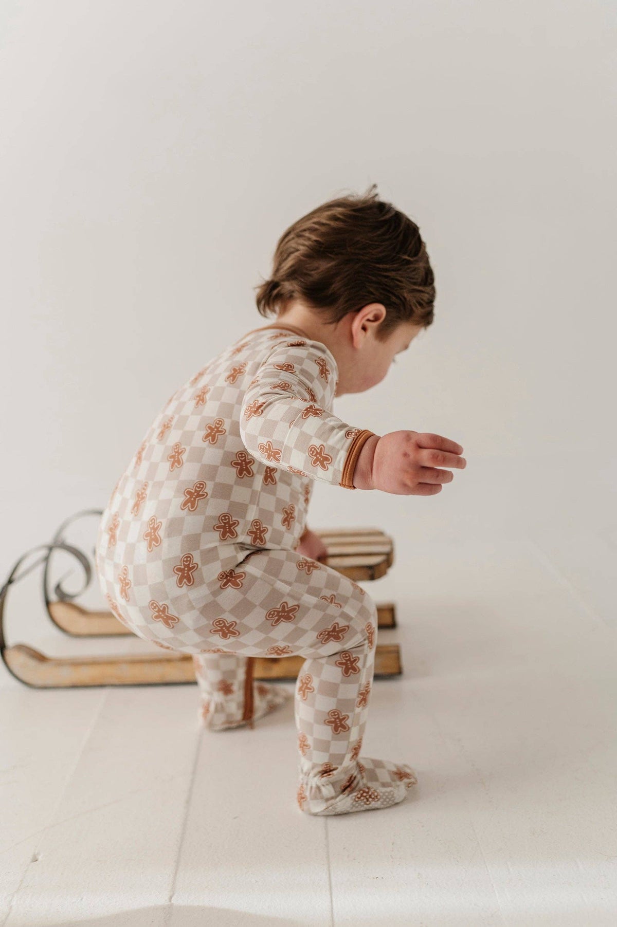 A baby wearing a long-sleeve footed pajama set with a beige and white checkerboard pattern and small brown gingerbread motifs, crouching on a white studio floor next to a wooden sled.