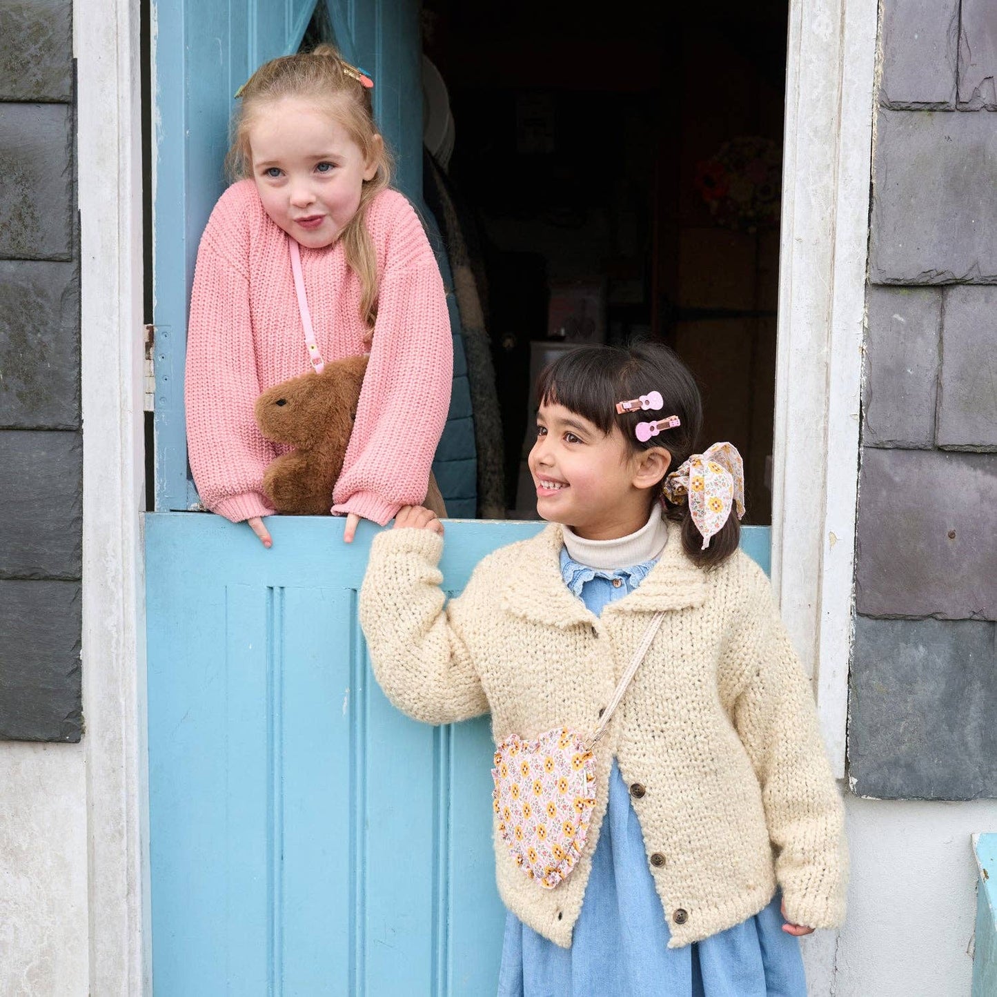 Two young girls wearing handmade knit cardigans and matching crossbody bags, posing on either side of a light blue wooden door. The girl on the left wears a pink knit cardigan with a brown teddy bear, and the girl on the right wears a cream knit cardigan with a blue dress and a floral patterned bag.