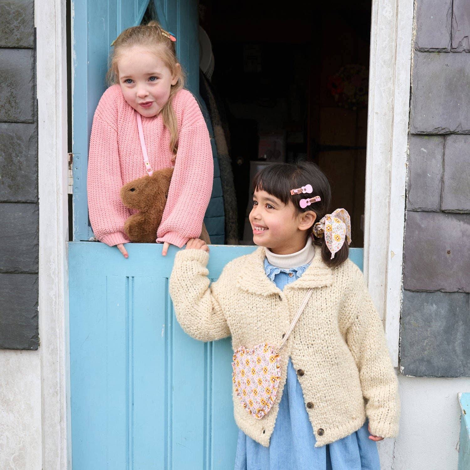 Two young girls wearing handmade knit cardigans and matching crossbody bags, posing on either side of a light blue wooden door. The girl on the left wears a pink knit cardigan with a brown teddy bear, and the girl on the right wears a cream knit cardigan with a blue dress and a floral patterned bag.