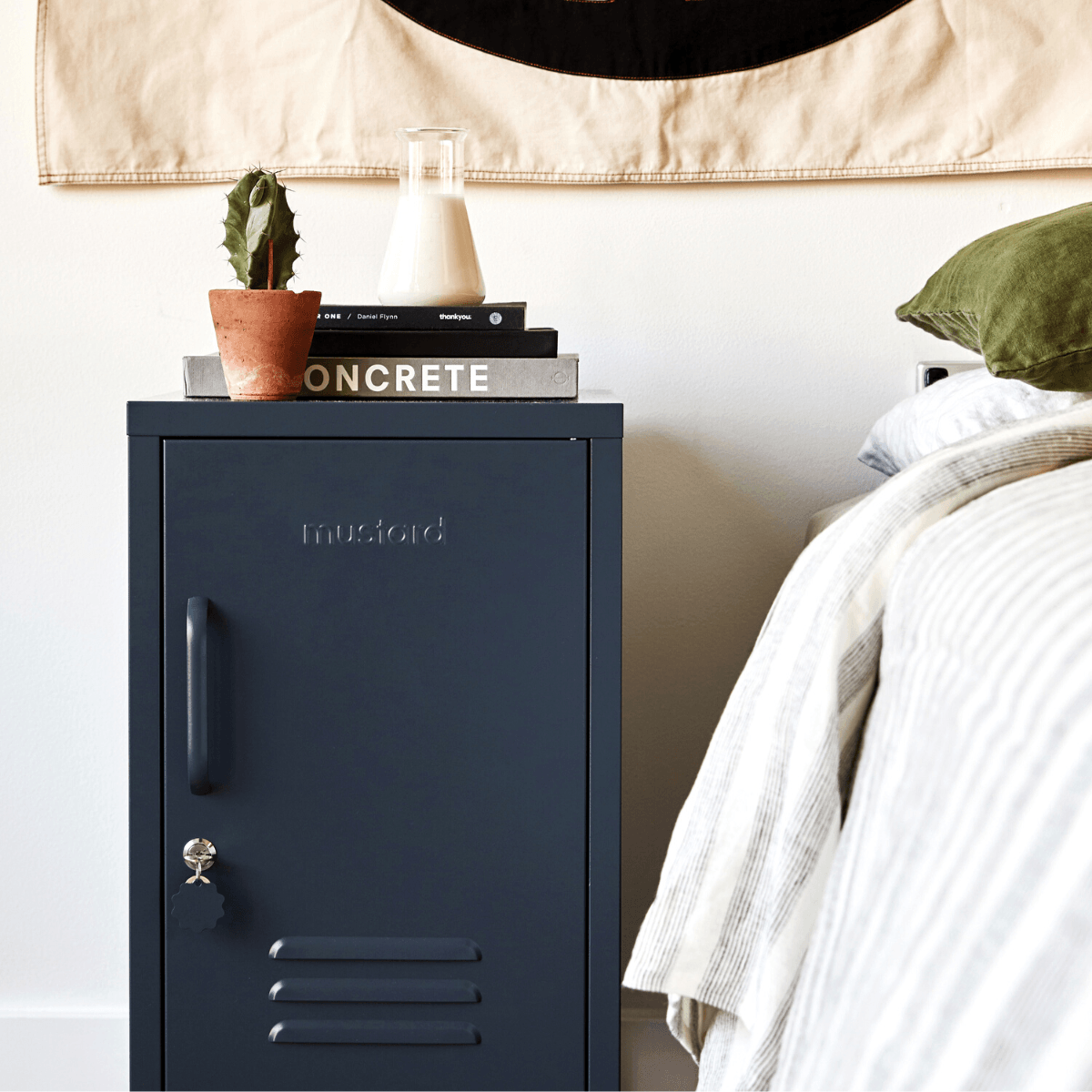 Gray cabinet with 'Mustard' branding next to a bed with green pillow and white bedding.