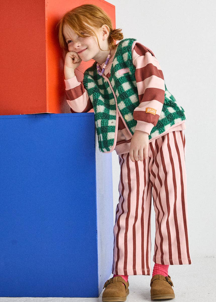 A young girl with red hair leans against a colorful geometric block wall. She is wearing a green and pink checkered zip-up vest over a pink and white striped long-sleeve top, paired with pink and white vertically striped pants, pink socks, and brown shoes.