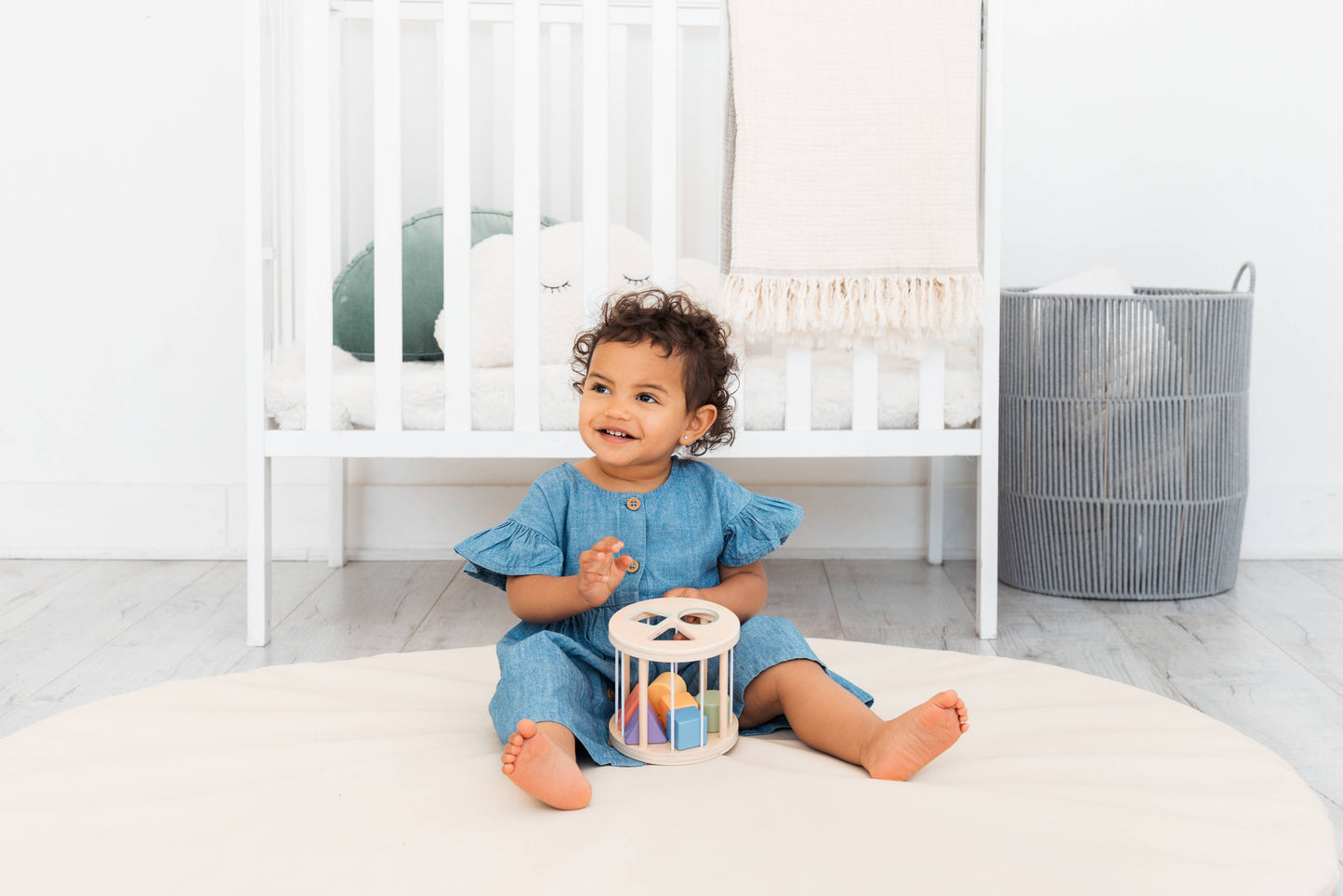 Child sitting on a white floor with a crib and basket in the background