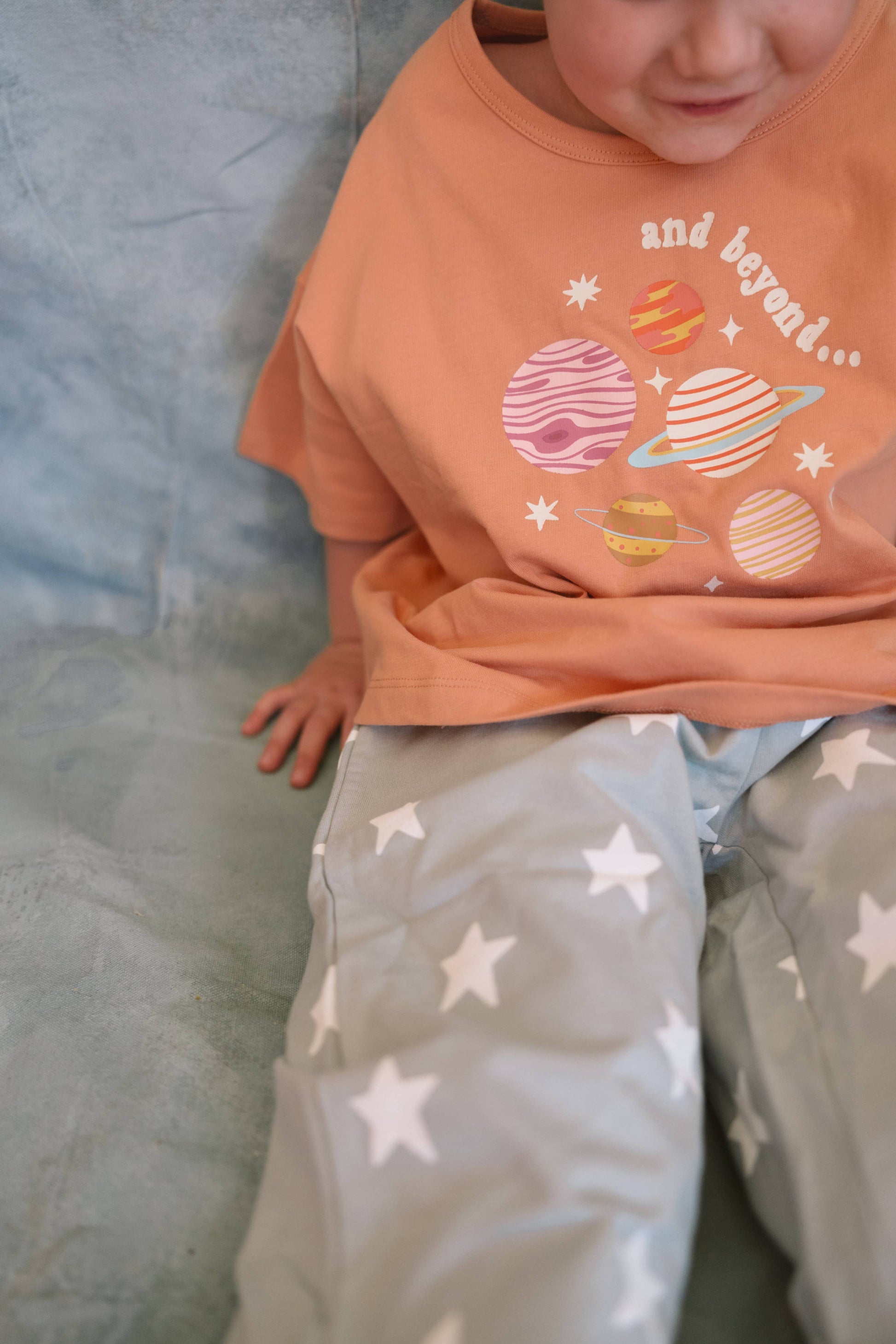 Child wearing an orange t-shirt with space-themed graphics and gray pants with star patterns.