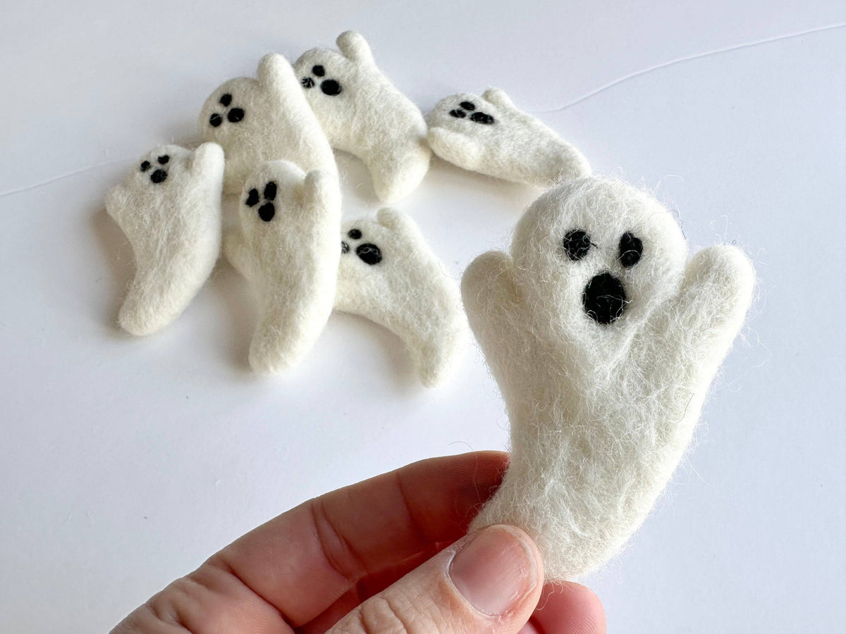A hand holds a white felted wool ghost, with several similar ghost finger puppets arranged in the background on a plain white surface.