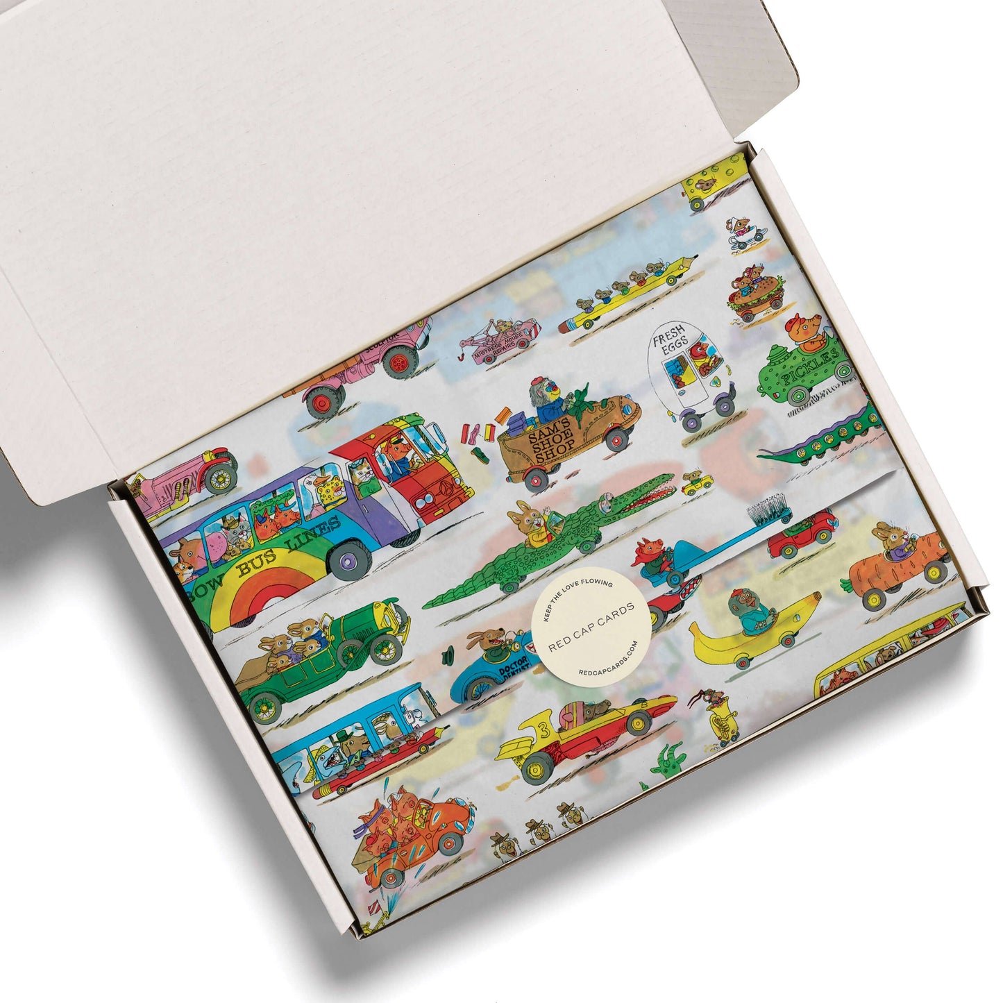 An open white cardboard gift box showing a colorful printed insert with illustrated vintage buses, cars, crocodiles, and small characters on a pale background. A round sticker in the center reads 'RED CAP CLOTHING'.